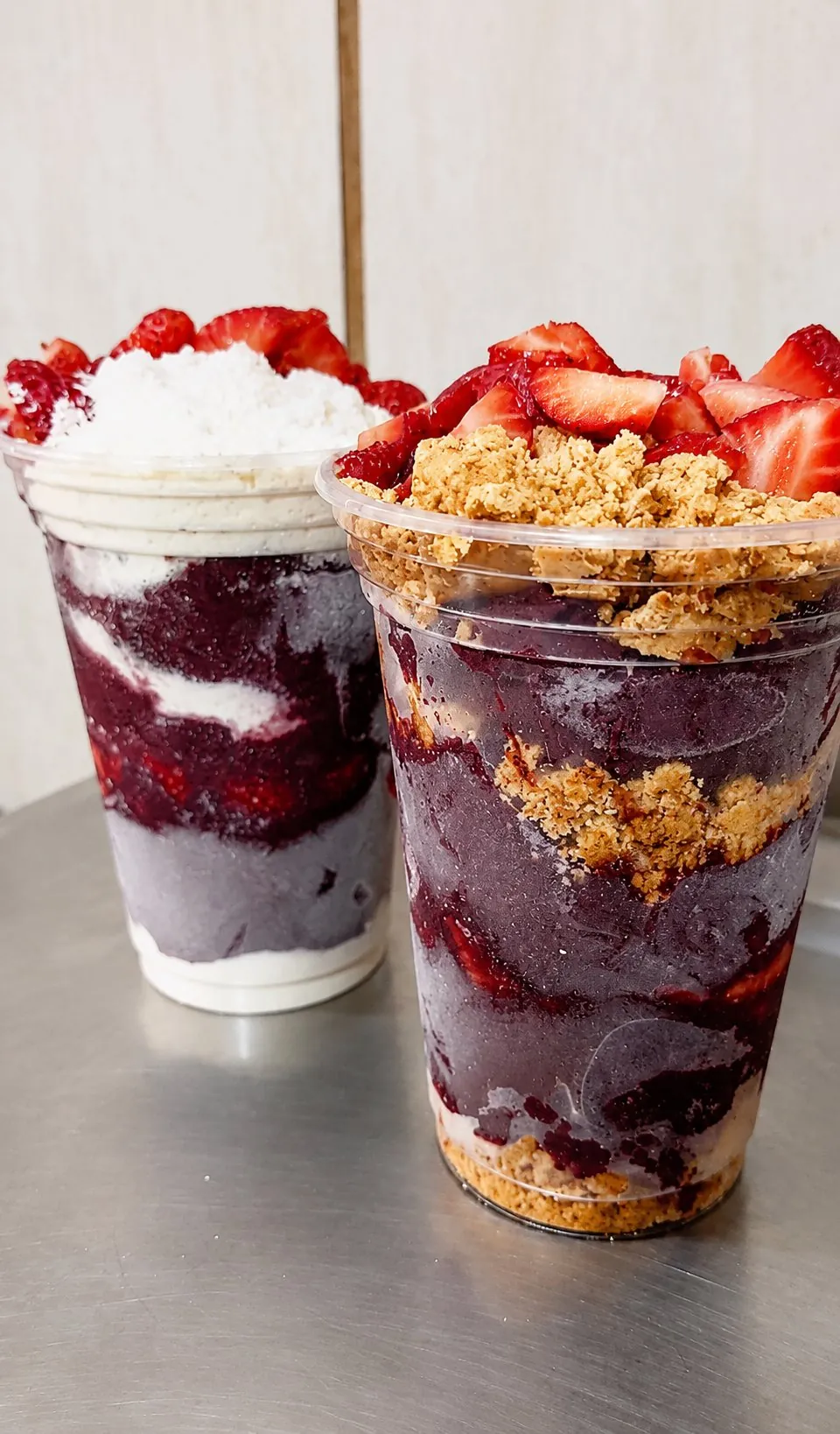 Dois copos de açaí montados com morango, granola e creme sobre balcão de inox