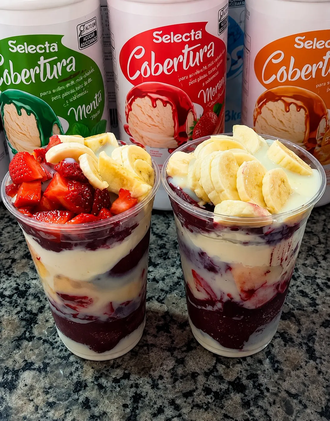 Dois copos de açaí com morango, banana e creme, com potes de cobertura ao fundo