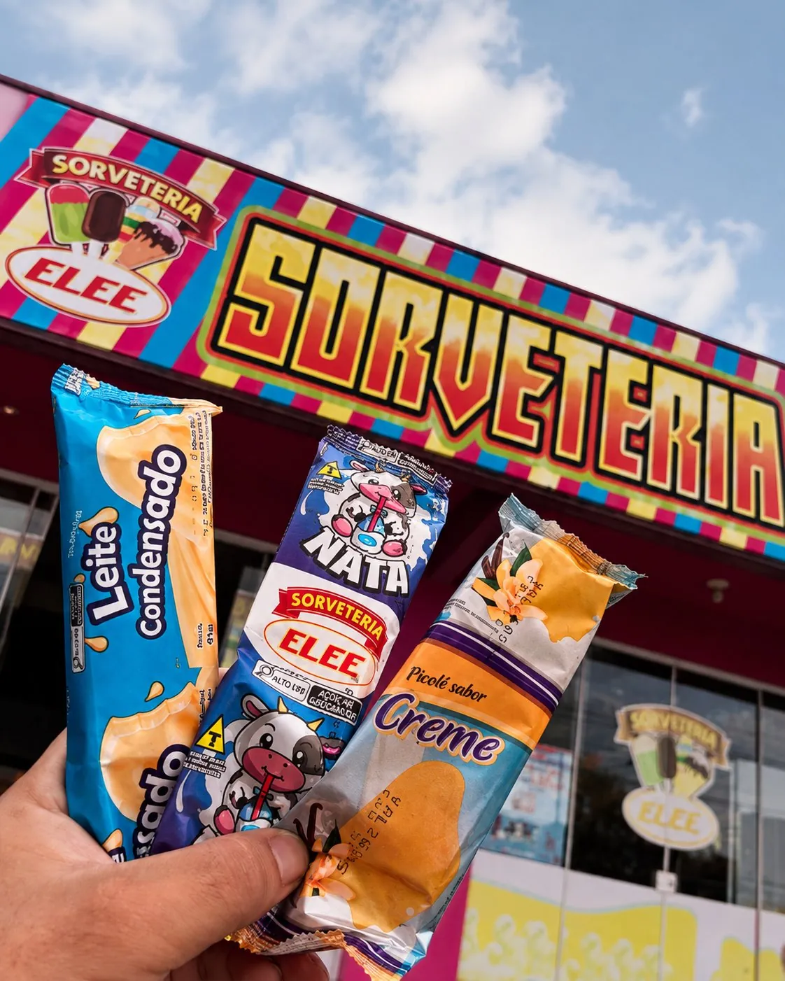 Três picolés da Sorveteria Elee segurados em frente à fachada colorida da loja
